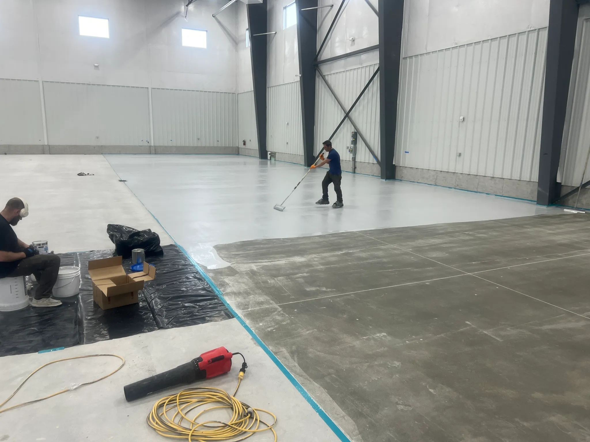 Worker applying gray epoxy coating to a large industrial concrete floor inside a high-ceiling warehouse